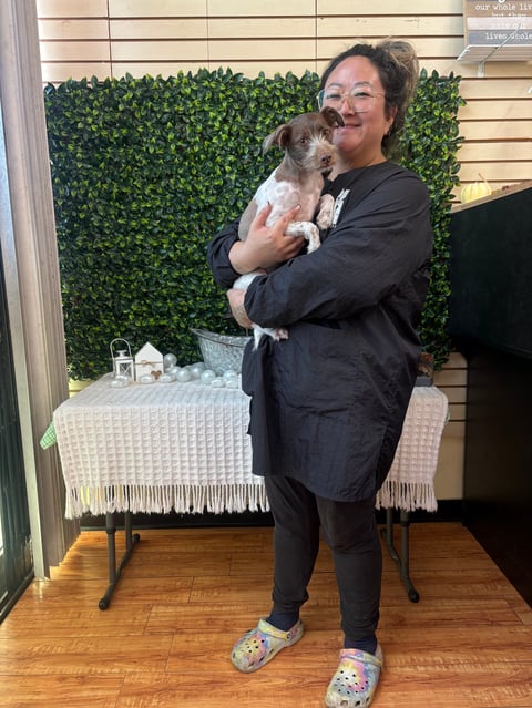 Woman in black jacket holding a small tan and white dog indoors with a decorative green plant wall backdrop behind her